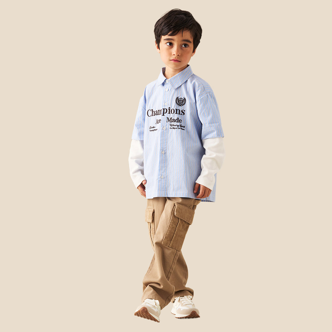 A child wearing wide-leg tan cargo pants with side flap pockets, styled with a light blue and white striped shirt and white sneakers against a solid beige background.