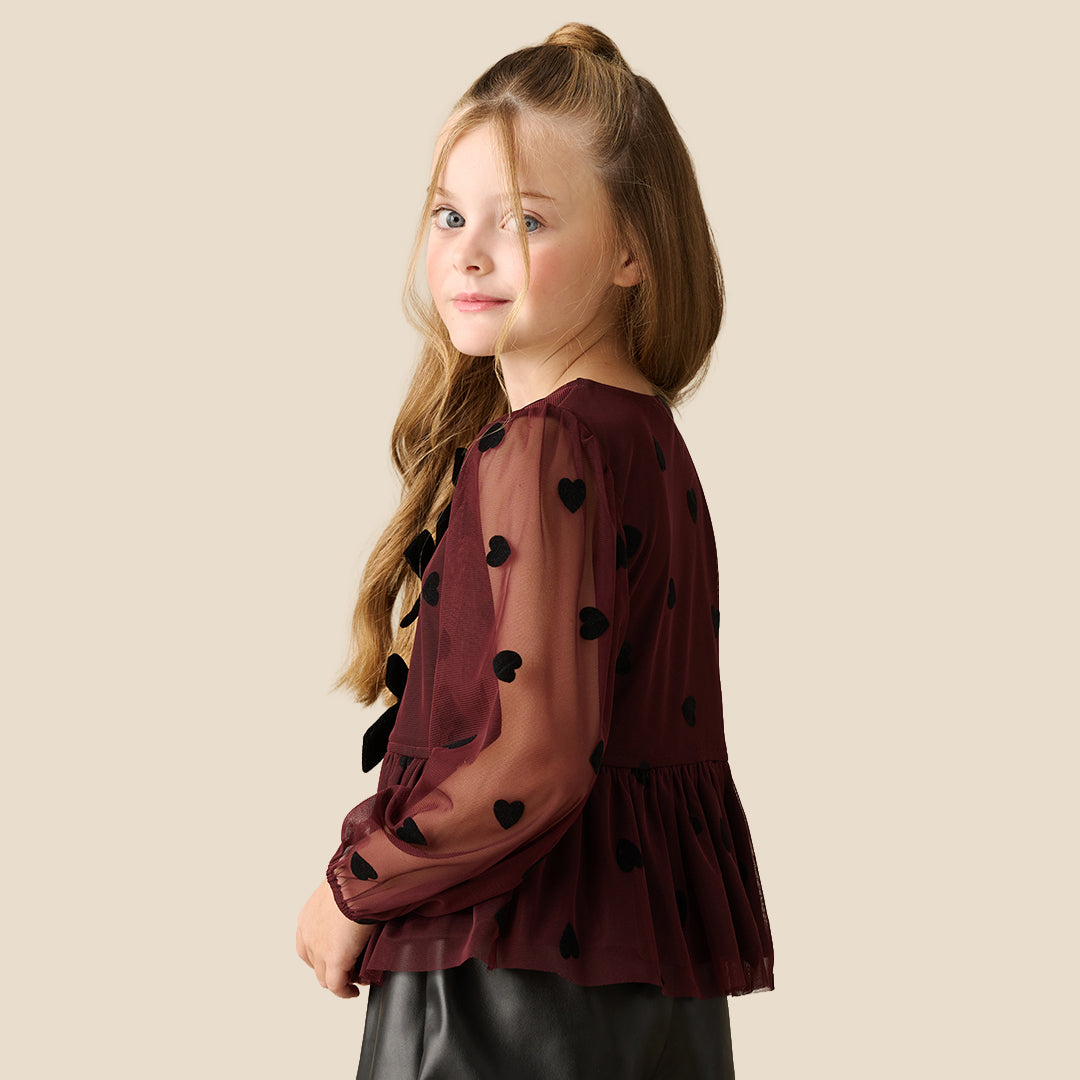 A young girl with blonde hair in a high ponytail is wearing a dark burgundy sheer long-sleeve blouse with black heart and bow details, paired with a black skirt, against a solid beige background.