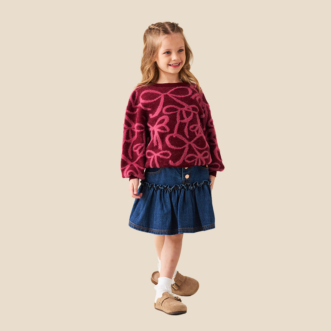 A young girl with blonde hair in a ponytail wearing a fuzzy dark red long-sleeve sweater with a pink bow pattern, paired with a blue denim skirt, against a solid beige background.