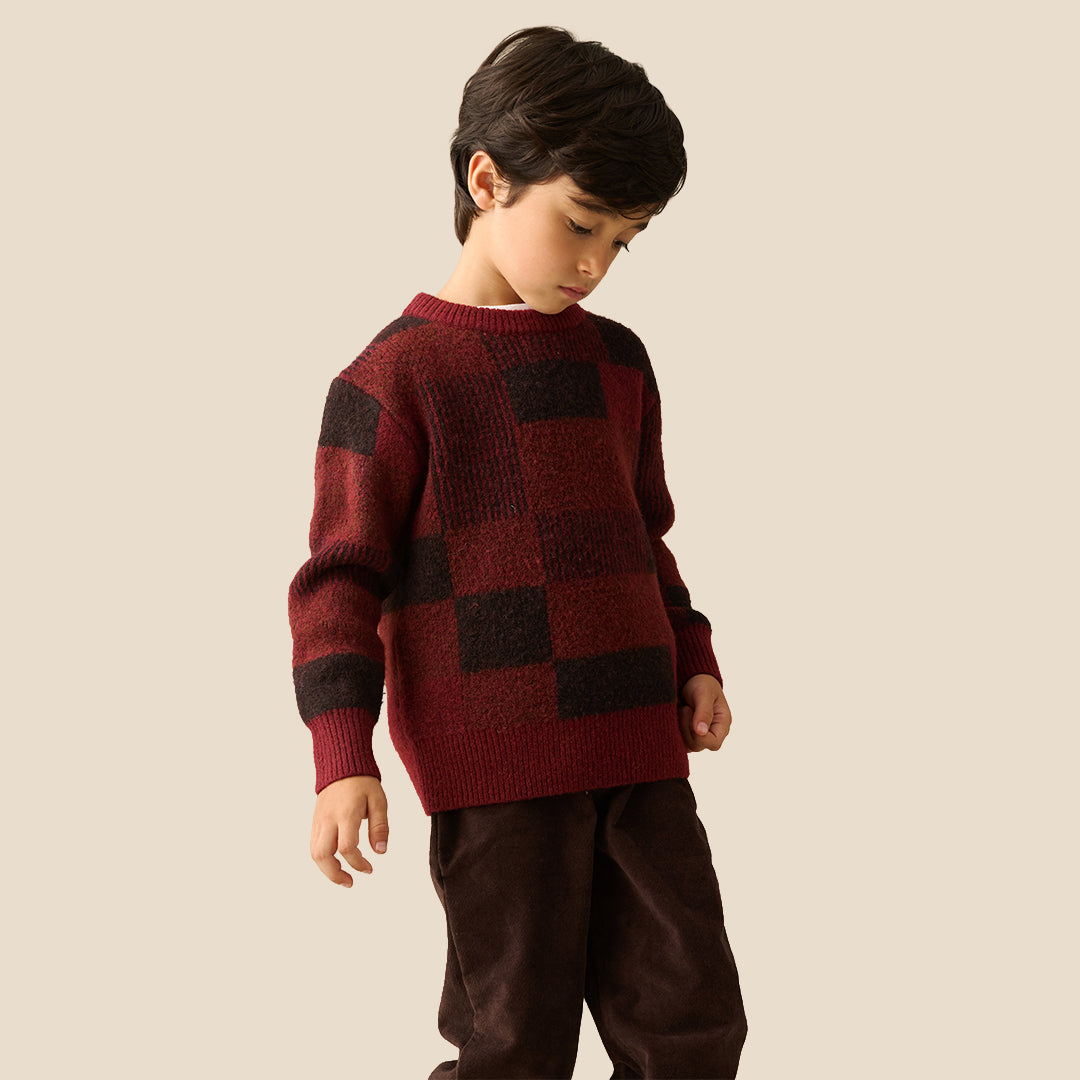A young boy wearing a textured knit sweater with a dark red and black checkered pattern, paired with dark brown pants, set against a solid light beige background.