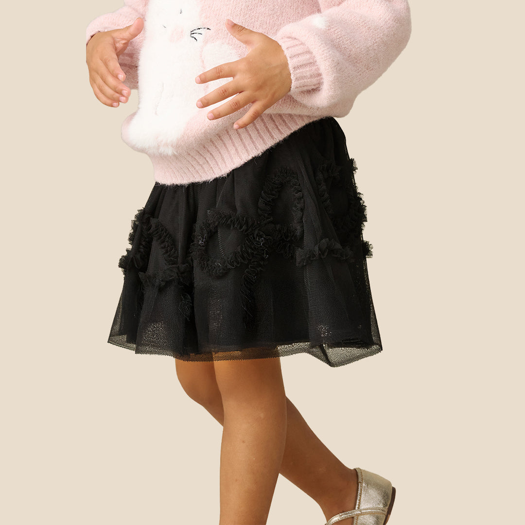 A child wearing a fluffy light pink sweater and a black tulle tutu skirt with ruffled trim, standing against a plain beige background.