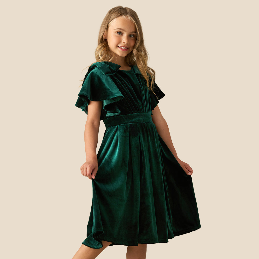 A young girl wearing a dark green velvet dress with ruffled short sleeves, a matching waist belt, and a pleated skirt, standing against a solid beige background.
