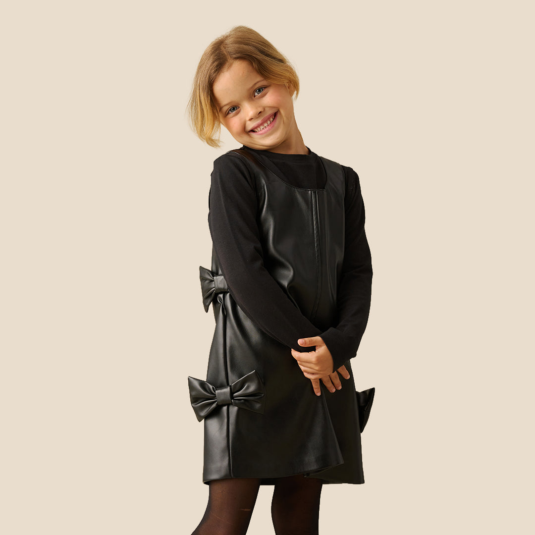 A young girl with blonde hair tied back is wearing a black faux-leather dress with long sleeves, a mock neck, and decorative bows on the sides, paired with black tights, against a solid beige background.