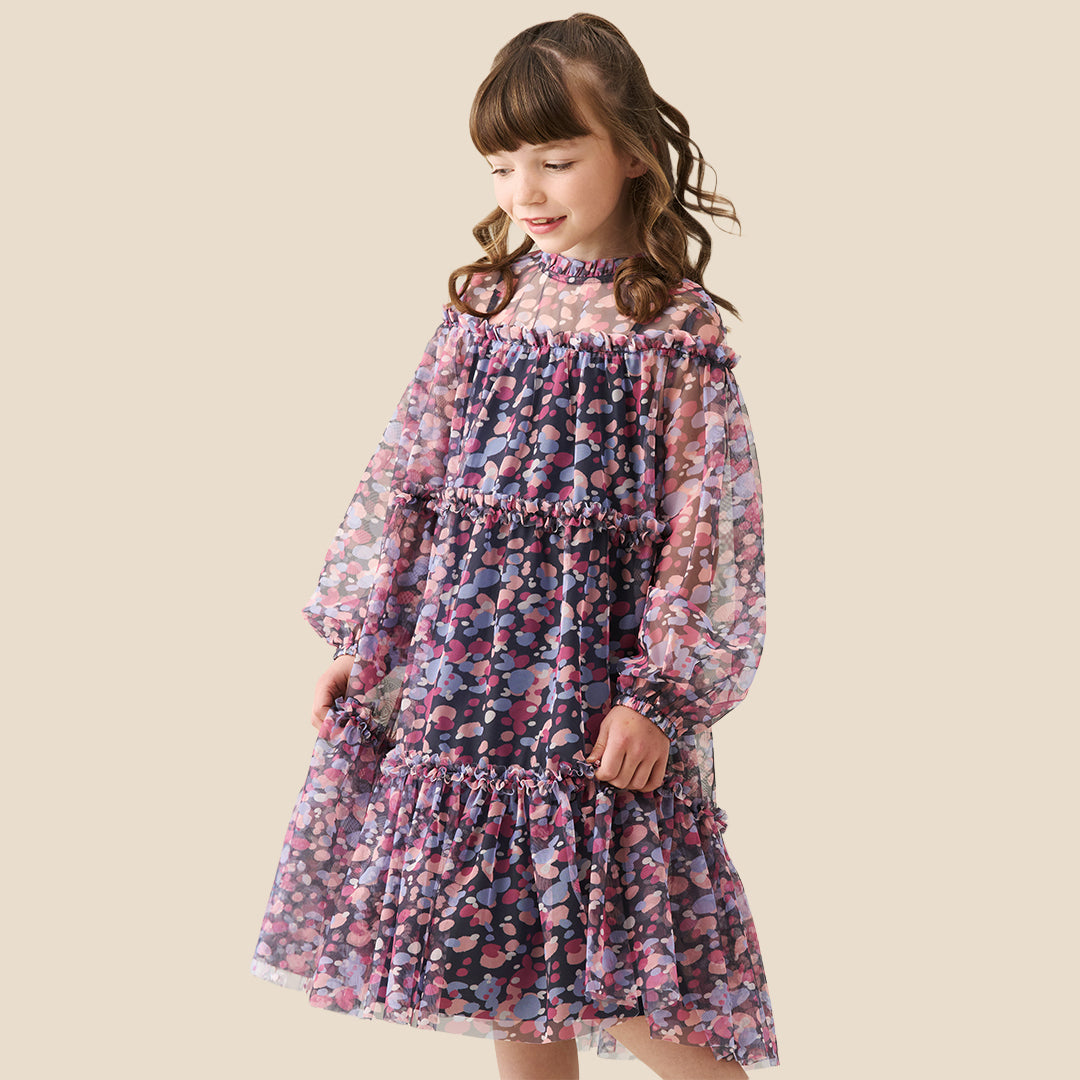A young girl with curly hair stands on a checkered porch wearing a long-sleeved tiered dress with a pink, purple, and blue floral pattern. She is posing behind a black wrought-iron fence.