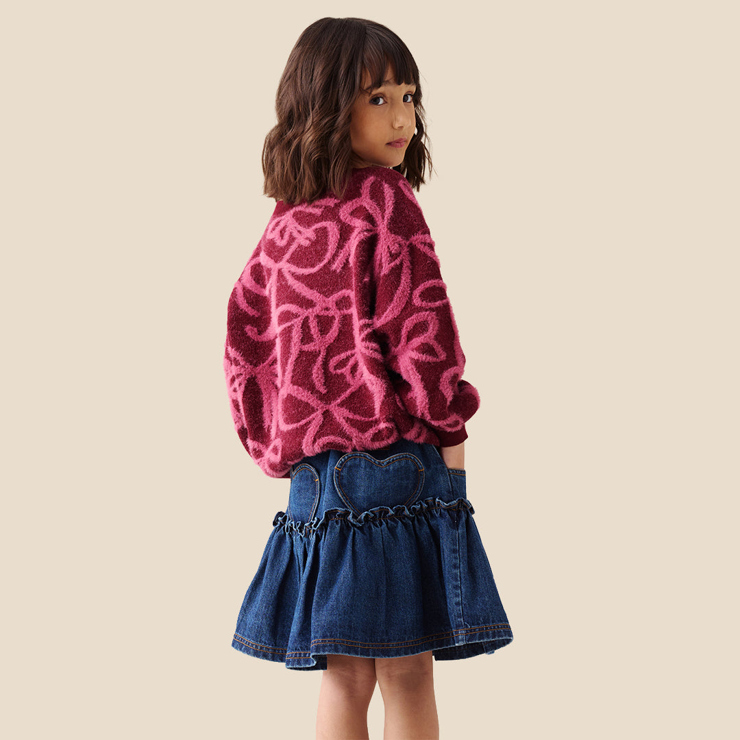 A young girl with blonde hair in a ponytail wearing a fuzzy dark red long-sleeve sweater with a pink bow pattern, paired with a blue denim skirt, against a solid beige background.