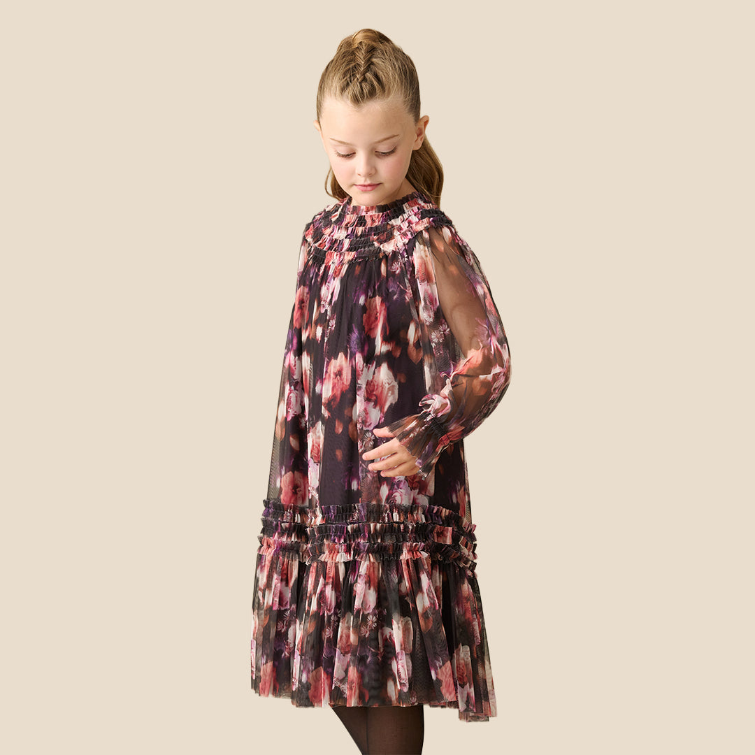 A young girl with her hair in a ponytail is wearing a long-sleeved, tiered tulle dress with a pink, red, and white floral pattern on a dark background, set against a solid beige studio backdrop.