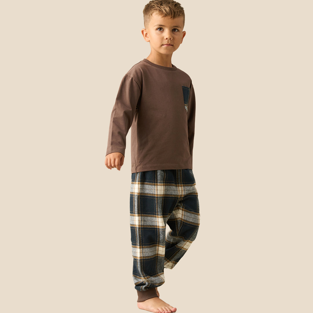 A young boy modeling a two-piece pajama set consisting of a brown long-sleeve shirt with a plaid chest pocket and matching plaid pants, set against a solid beige background.