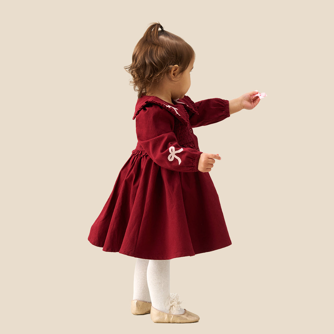 A toddler girl wearing a burgundy corduroy dress with a large white ruffled collar decorated with bows, white tights, and beige ballet flats, standing against a solid beige background.