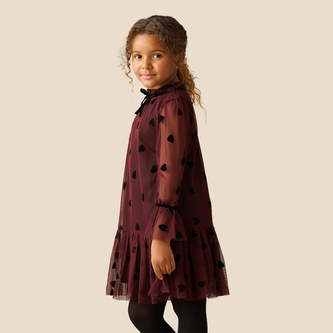 A young girl with curly brown hair wearing a burgundy sheer mesh dress with black heart patterns and a ruffled tulle hem, standing against a solid beige background.