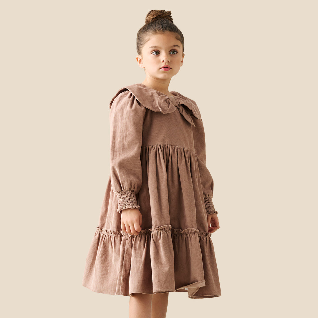 A young girl with her hair in a bun wears a brown corduroy dress with a large ruffled collar and bow, standing against a solid beige background.