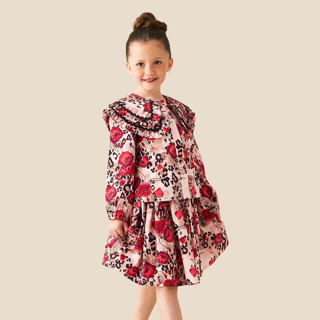 A young girl with her hair in a bun stands wearing a long-sleeve dress with a red rose and black leopard print pattern on a light pink background, set against a solid beige studio backdrop.
