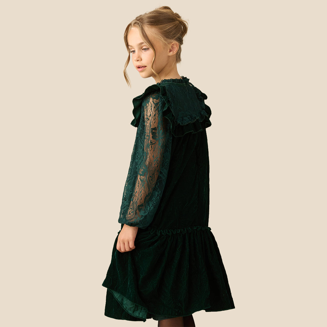 A young girl wearing a dark forest green velvet dress with lace sleeves and a ruffled high neckline, photographed against a solid beige background.