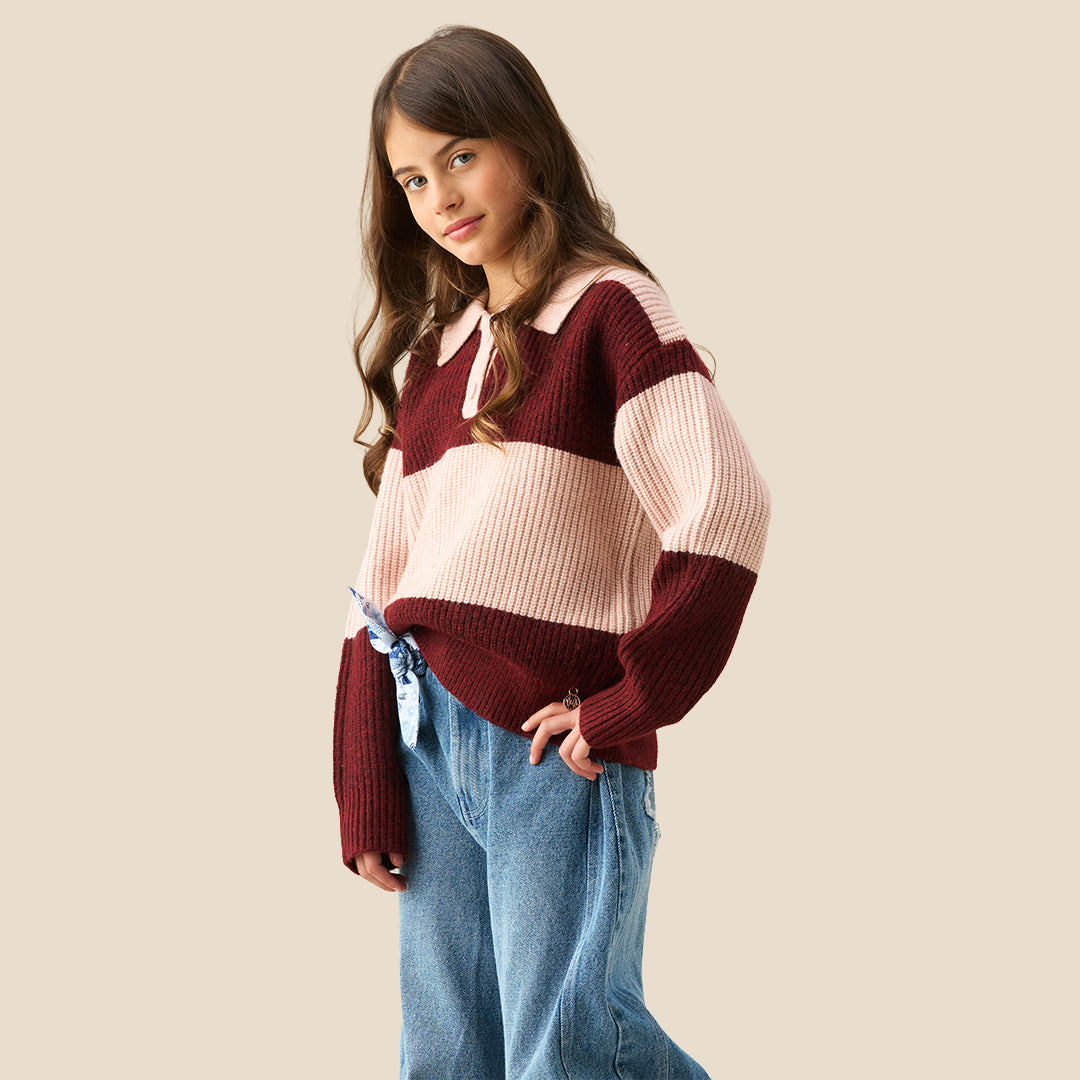 A young girl modeling a ribbed knit polo sweater with horizontal stripes in burgundy and light pink, paired with wide-leg blue jeans against a solid beige background.