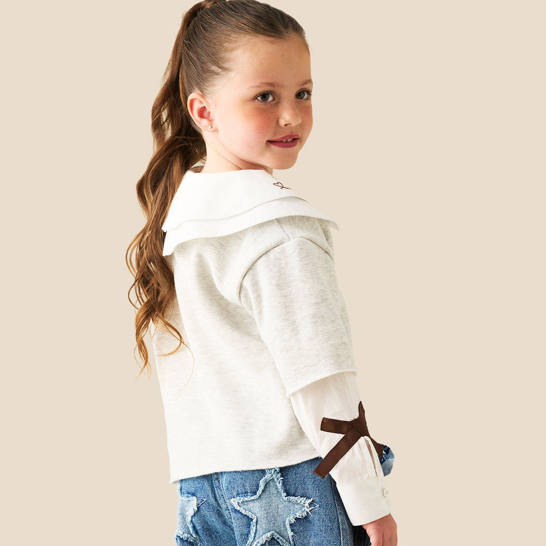 A young girl with her hair in a ponytail models a light gray long-sleeve top featuring a large white collar with brown embroidered details and matching brown accents on the sleeves, paired with blue jeans against a solid beige background.