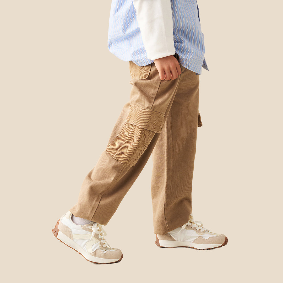 A child wearing wide-leg tan cargo pants with side flap pockets, styled with a light blue and white striped shirt and white sneakers against a solid beige background.