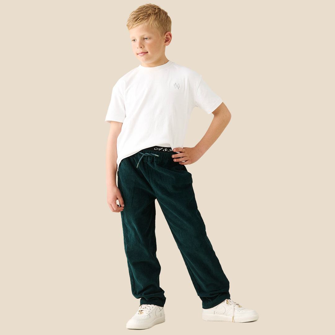 A young boy modeling a white crewneck t-shirt and dark green corduroy pants, standing against a solid beige background.