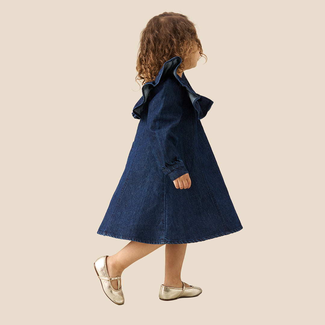 A young child wearing a dark blue denim dress with dramatic ruffled shoulders, standing against a solid beige background.