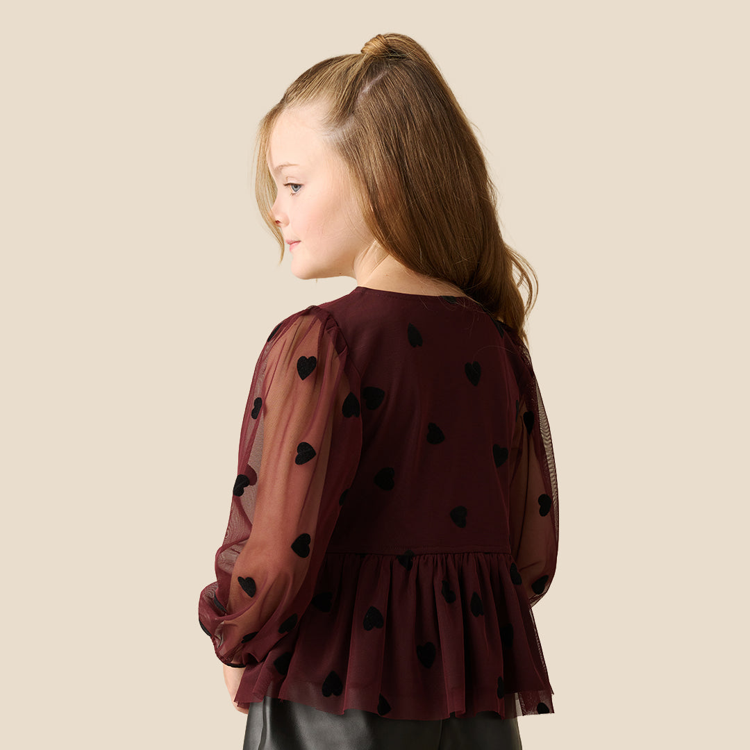 A young girl with blonde hair in a high ponytail is wearing a dark burgundy sheer long-sleeve blouse with black heart and bow details, paired with a black skirt, against a solid beige background.