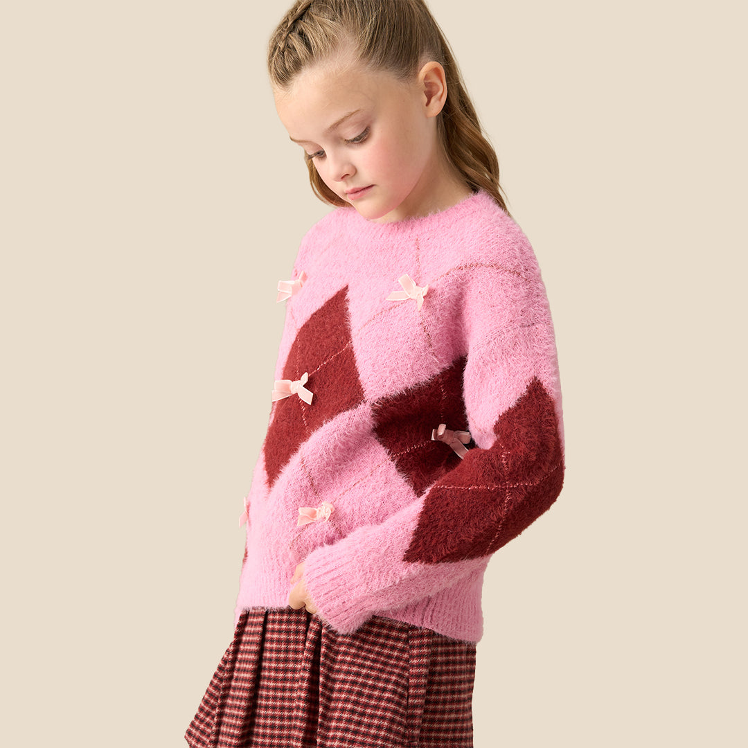A young girl modeling a pink and dark red argyle sweater with small bow details, paired with a red and black gingham pleated skirt, against a solid beige background.