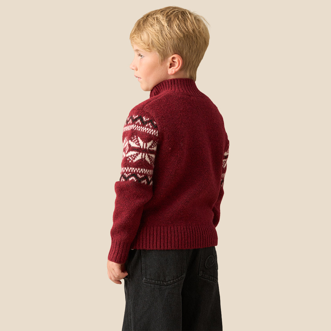 A young boy smiling and wearing a burgundy knit quarter-zip sweater with a white and dark red Fair Isle snowflake pattern across the chest and shoulders, set against a solid beige background.