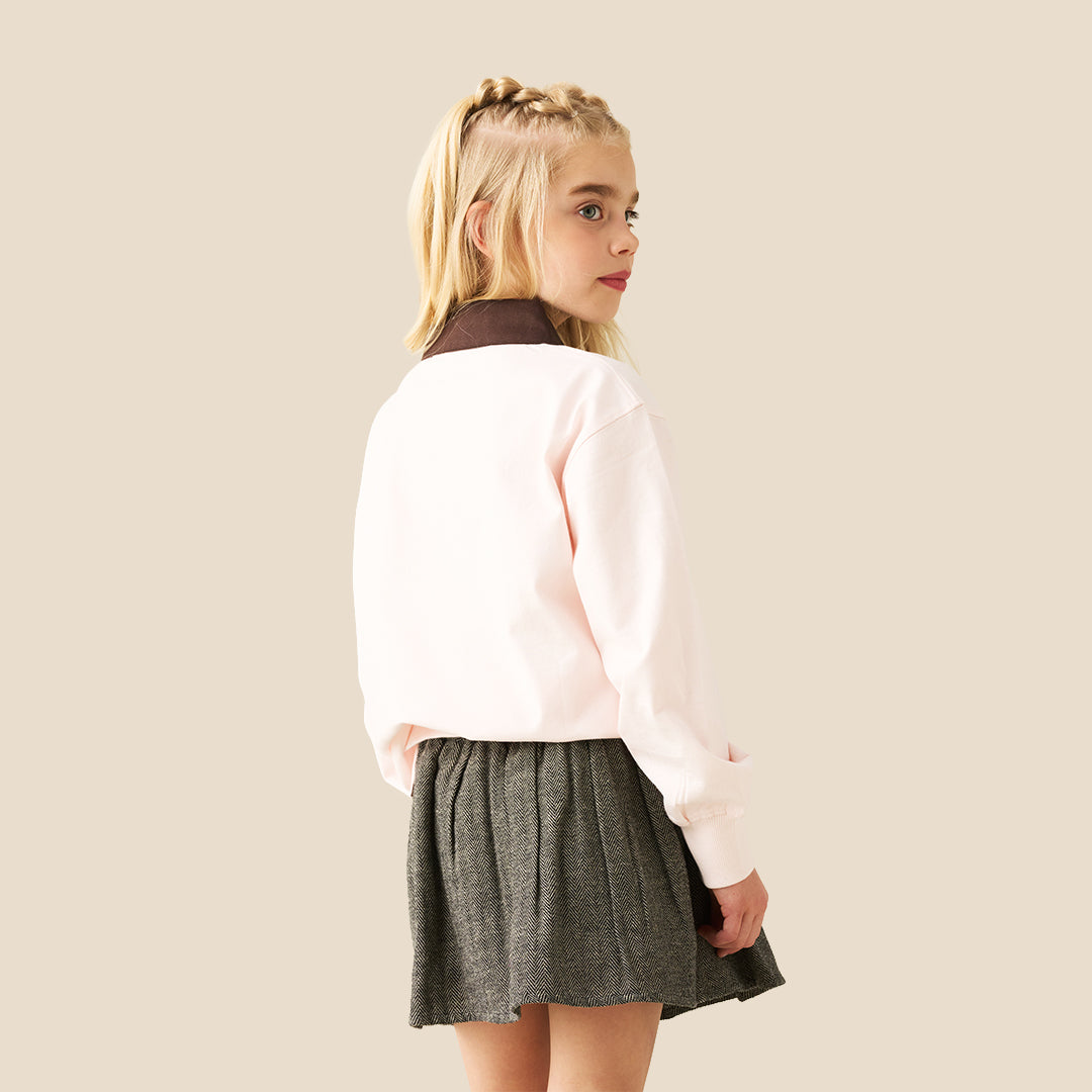 A model wearing a light pink sweatshirt with brown cursive text and a dark olive green pleated tweed mini skirt, standing against a solid beige background.