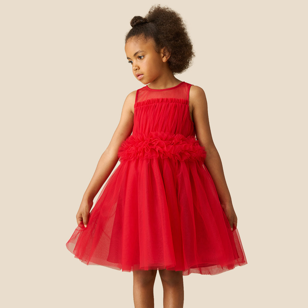A young girl with curly brown hair wearing a sleeveless bright red tulle dress with a ruffled waistband, standing against a solid beige background.