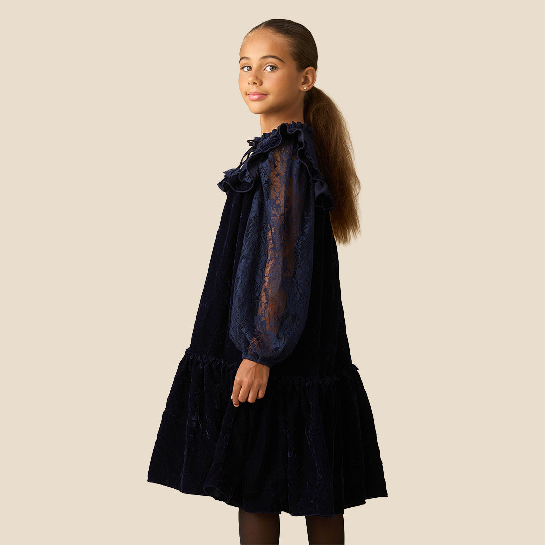 A young girl with her hair in a ponytail models a dark navy blue dress. The dress features a velvet-like texture, a ruffled collar, sheer lace long sleeves, and a tiered skirt, set against a solid beige background.