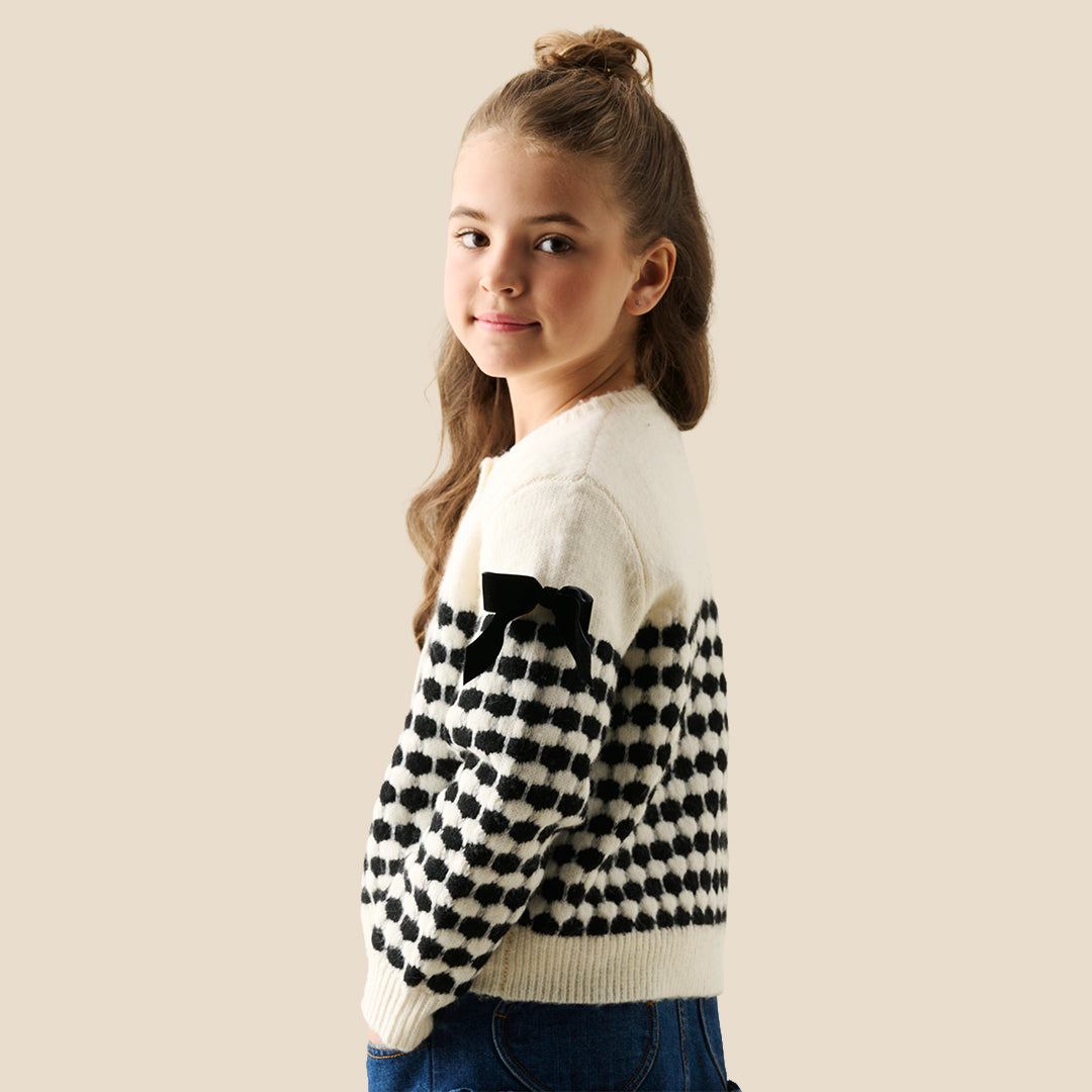 A young girl with her hair in a bun wears a cream knit cardigan with black polka-dot patterned sleeves and black ribbon bows on the shoulders, paired with a dark blue ruffled skirt, against a solid beige background.
