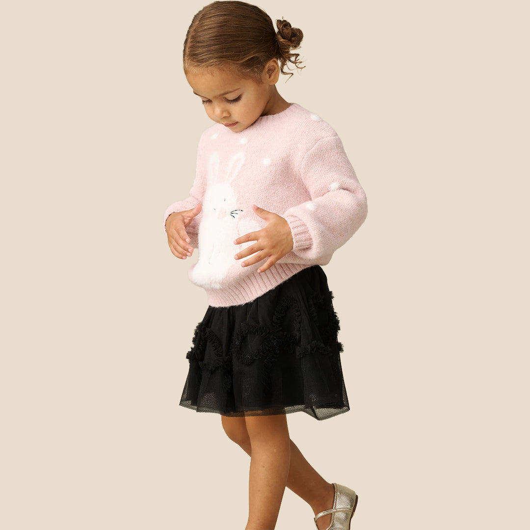 A child wearing a fluffy light pink sweater and a black tulle tutu skirt with ruffled trim, standing against a plain beige background.