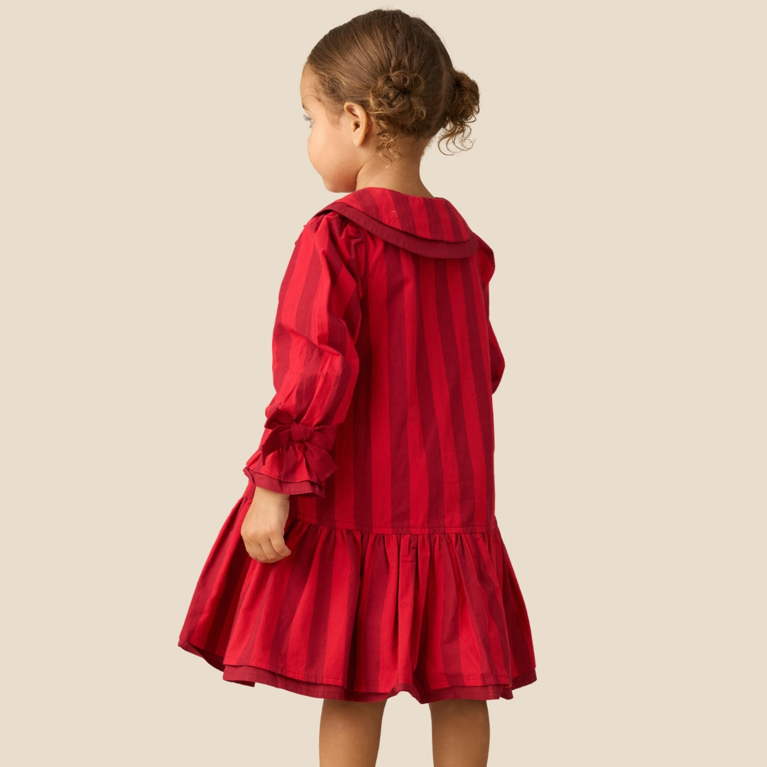 A young girl with her hair in a bun is shown from the back, wearing a red and dark red vertically striped long-sleeve dress with a ruffled collar and a flared skirt, set against a solid beige background.