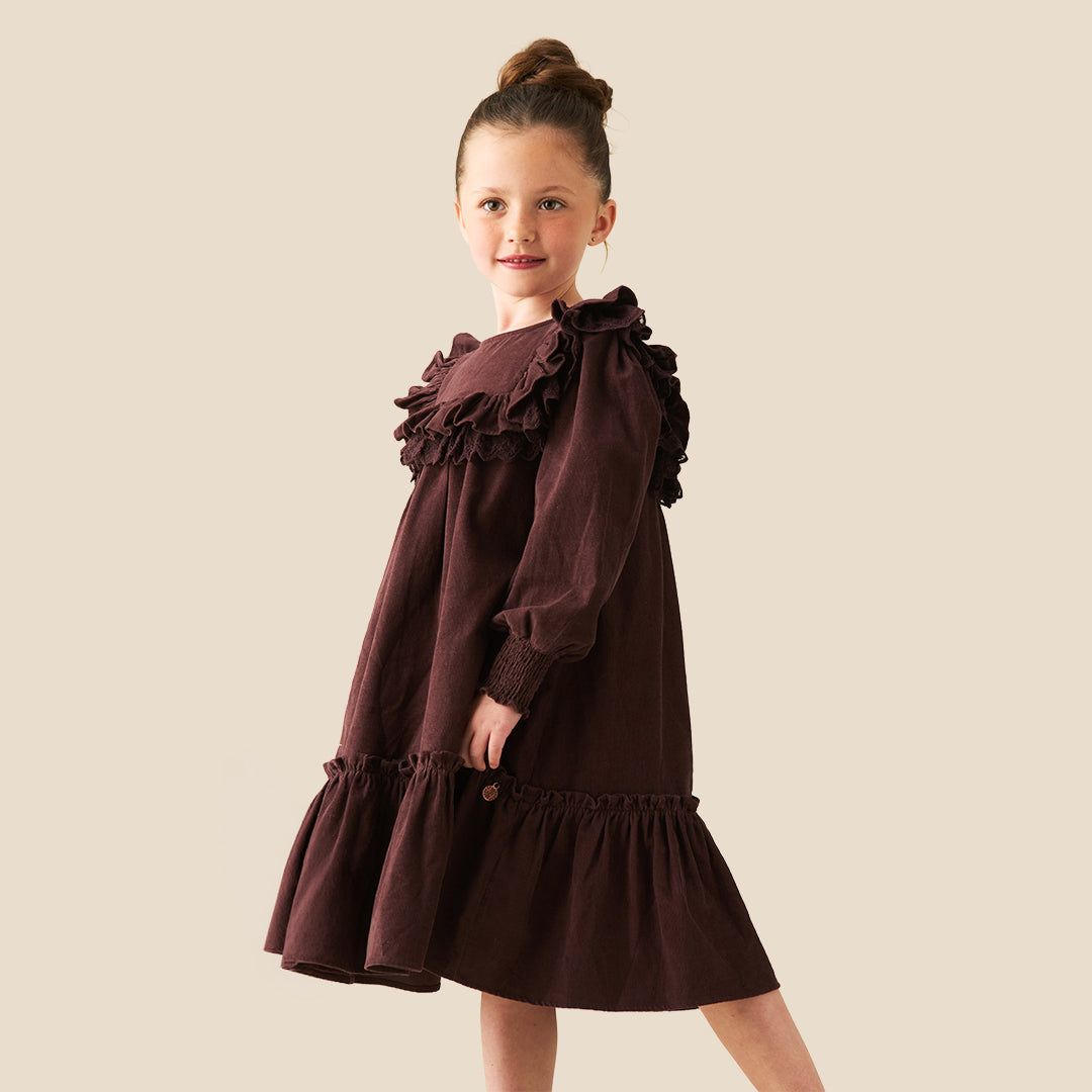 A young girl with her hair in a high bun models a dark brown long-sleeve dress with ruffled details, set against a solid light beige background.