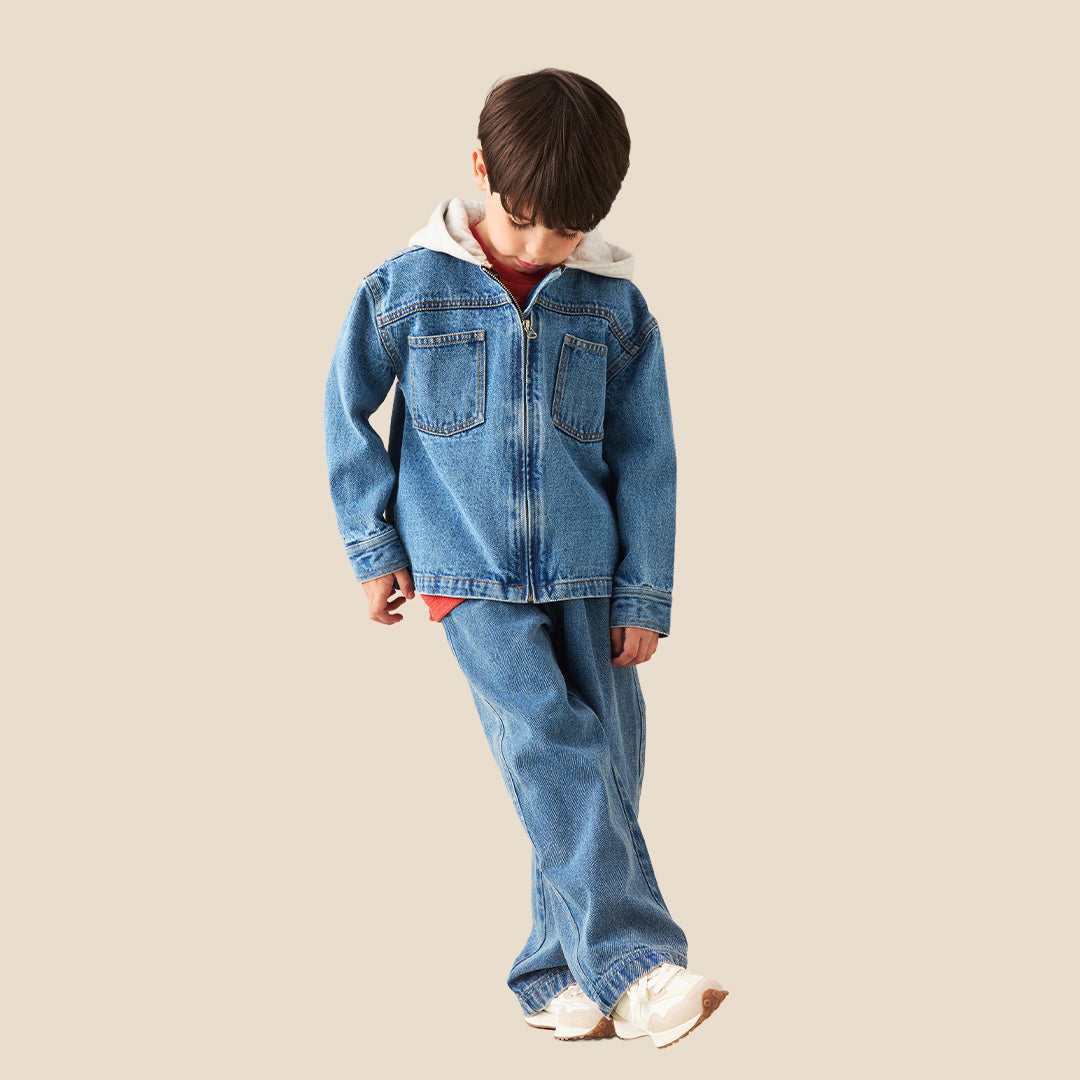 A young boy wearing a light blue denim zip-up jacket with a white hood lining and matching blue jeans, standing against a solid beige background.