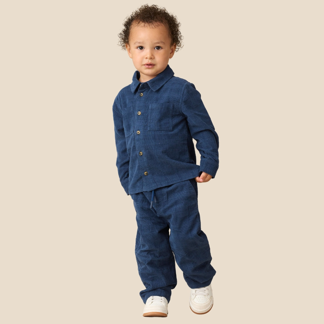 A toddler standing against a plain beige background, wearing a matching dark blue corduroy button-up shirt and pants set with white sneakers.