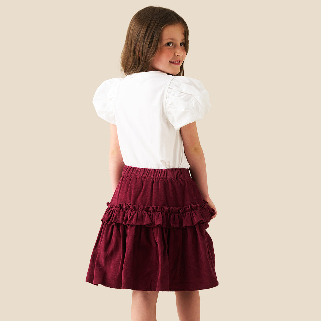 A child wearing a tiered burgundy skirt with ruffles and a white puff-sleeve top featuring a pink plaid bow graphic, photographed against a plain beige background.