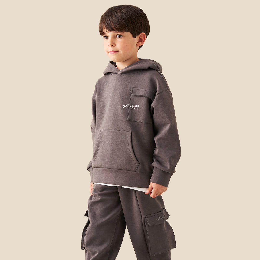 A young boy wearing a dark gray hoodie with a small white graphic on the chest and matching dark gray cargo pants, standing against a solid beige background.
