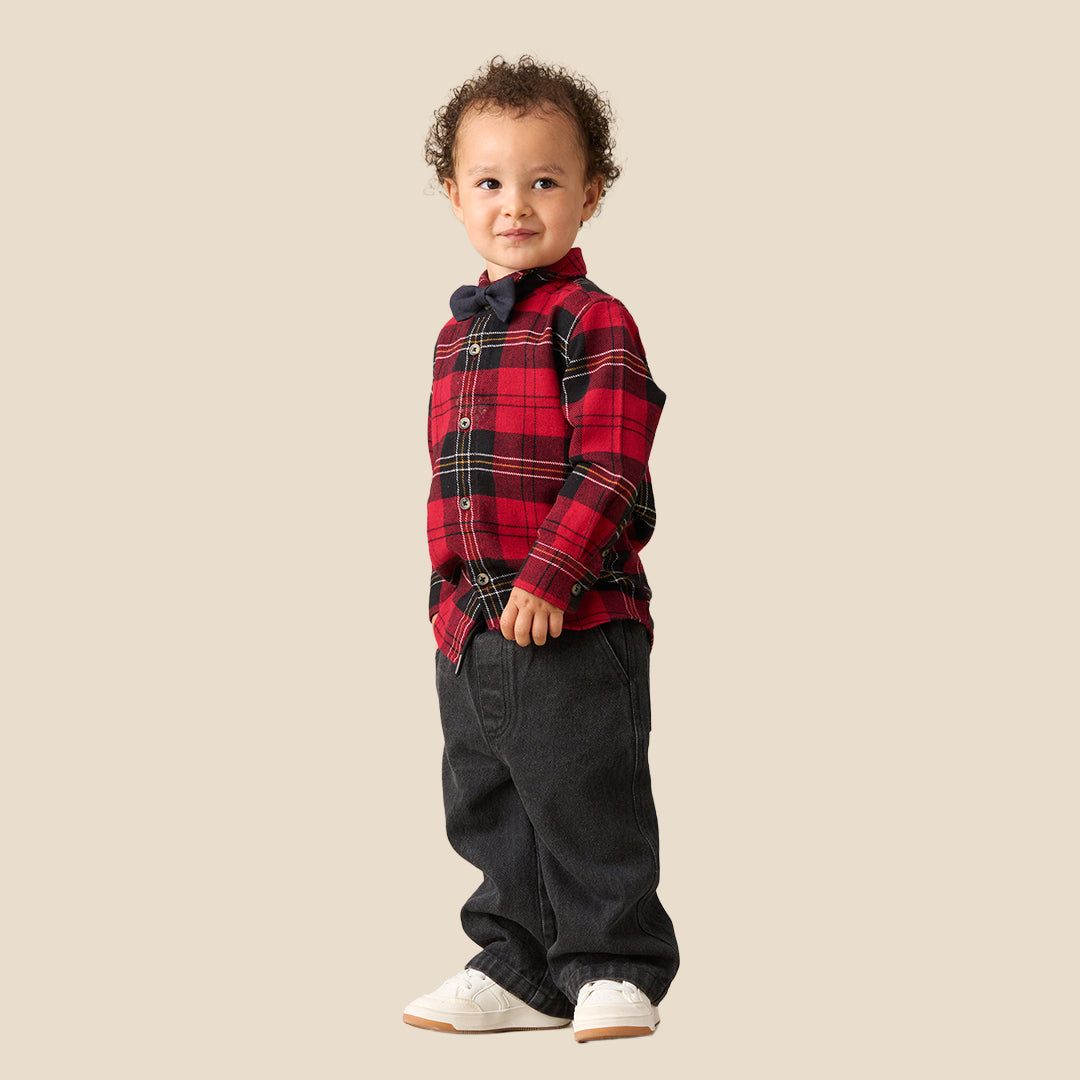A toddler standing against a plain beige background, wearing a red and black plaid long-sleeve shirt with a matching dark bow tie, dark pants, and white sneakers.