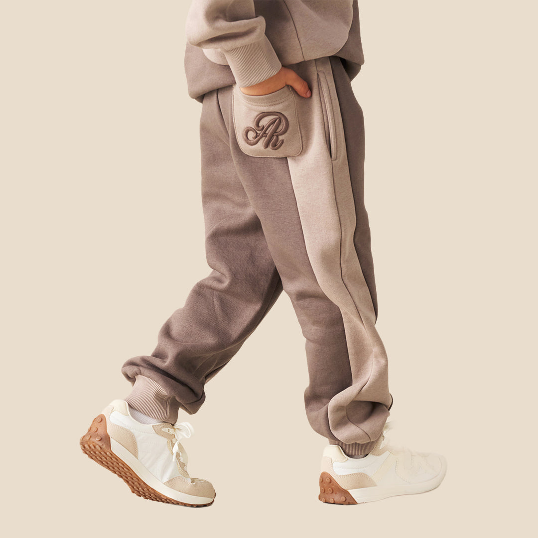 A person wearing loose taupe-brown jogger sweatpants with a white drawstring, styled with a beige and cream sweatshirt and white sneakers, against a solid beige background.