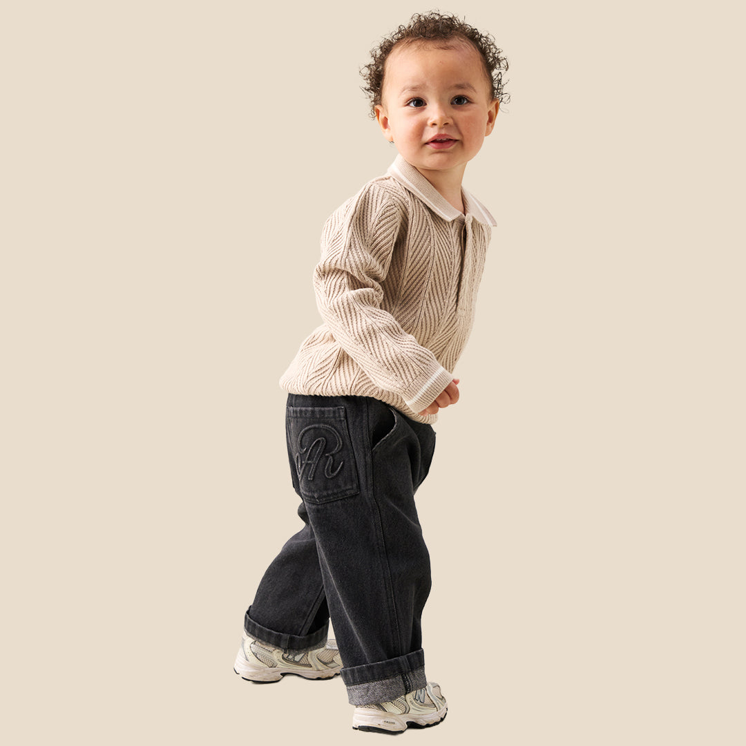 A toddler with curly brown hair wearing a cream-colored cable-knit sweater with a white Peter Pan collar, standing against a solid beige background.