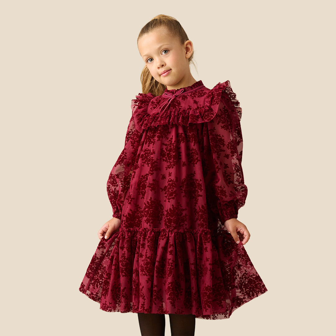 A young girl with her hair in a ponytail models a long-sleeved burgundy dress with a floral lace overlay, ruffled collar, and tiered skirt, set against a solid beige background.