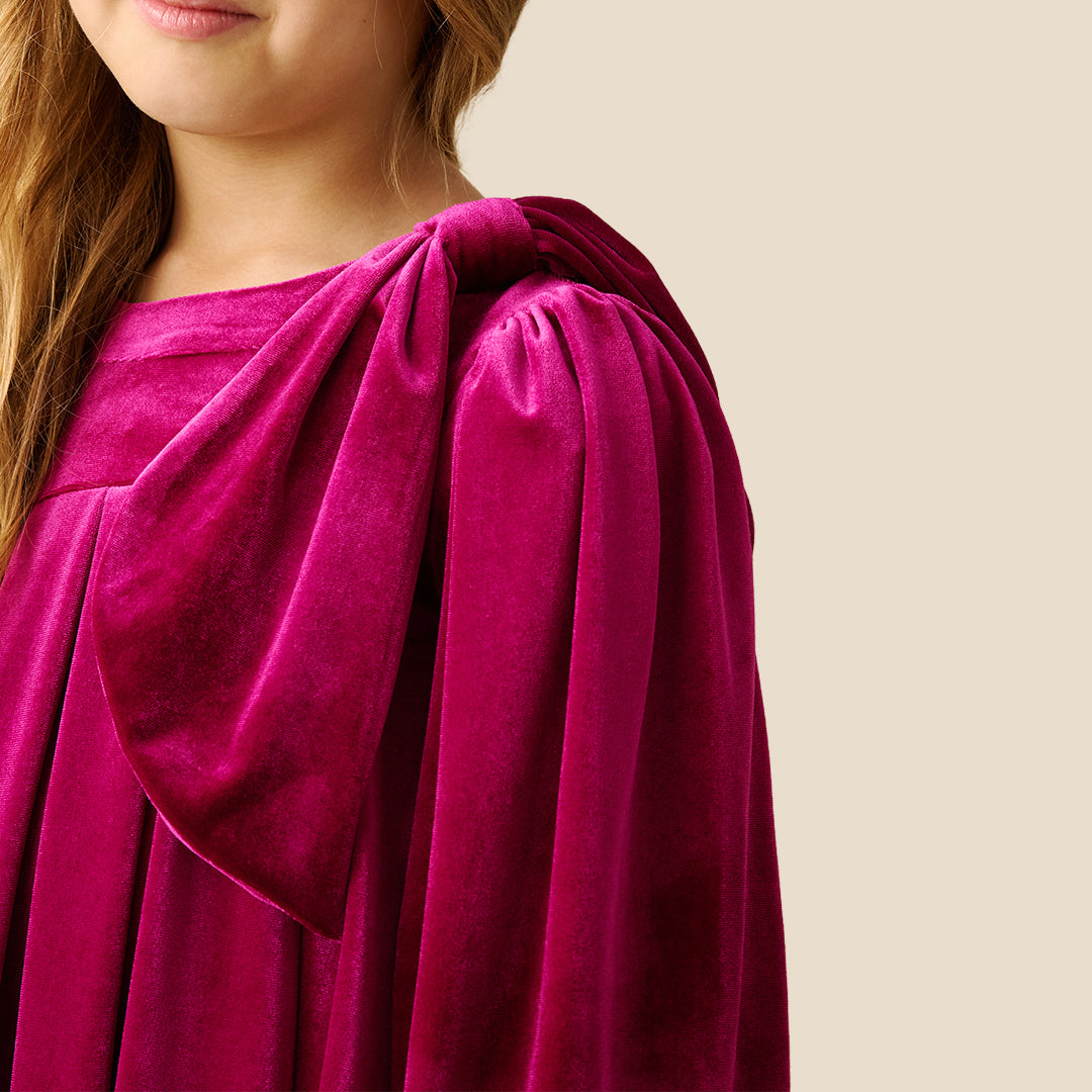 A young girl with blonde hair wearing a long-sleeved magenta velvet dress with a gathered bow detail on the chest, standing against a solid beige background.