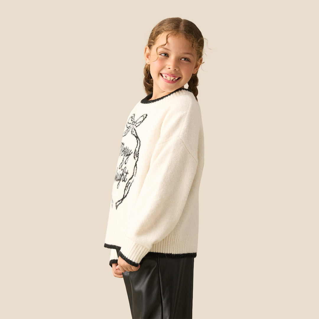 A young girl smiling while wearing a cream-colored knit sweater with a black 'Merry & Bright' ornament graphic and black trim, paired with black faux leather pants, against a solid beige background.