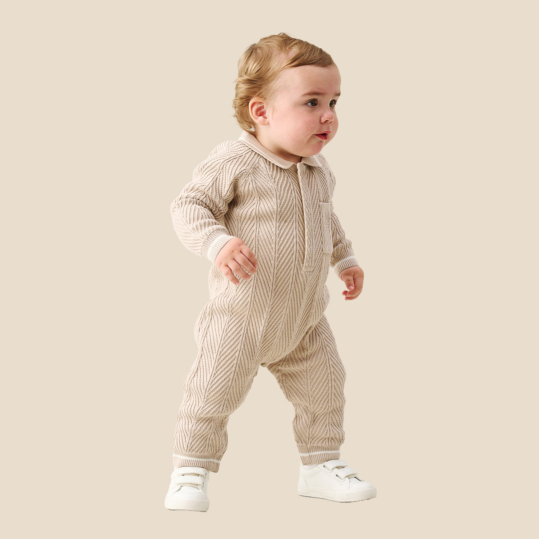 A baby wearing a beige and white textured knit one-piece jumpsuit with white shoes, standing against a solid beige background.