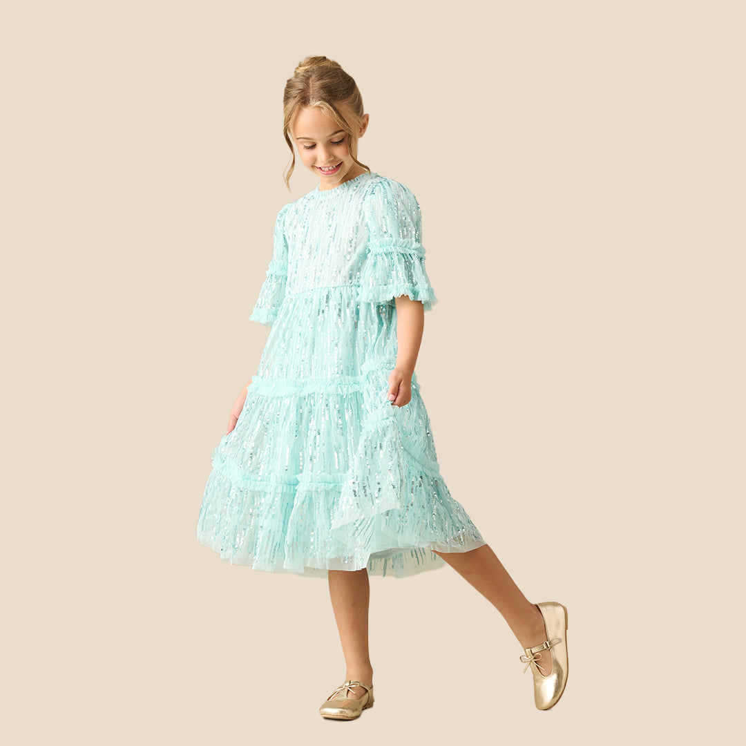 A young girl with a long brown ponytail wearing a light mint green sequined tiered dress, standing against a solid beige background.