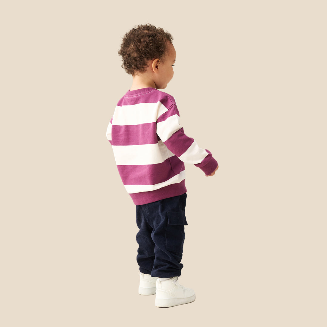 A toddler standing against a solid beige background, wearing a purple and white horizontally striped long-sleeve sweatshirt, dark navy pants, and white sneakers.