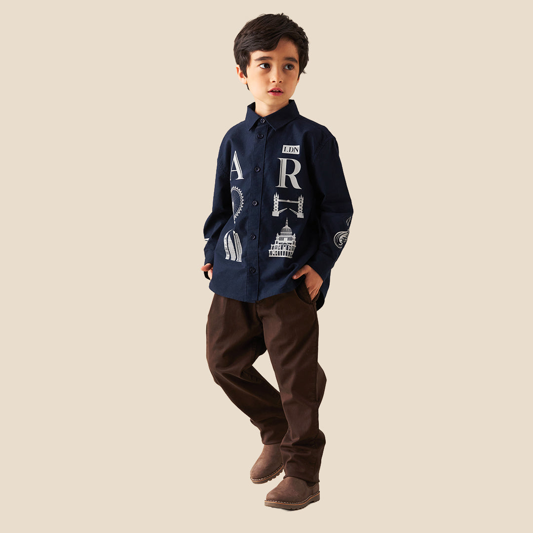 A child wearing dark brown trousers and a navy blue button-up shirt with white graphic prints, standing against a solid beige background.