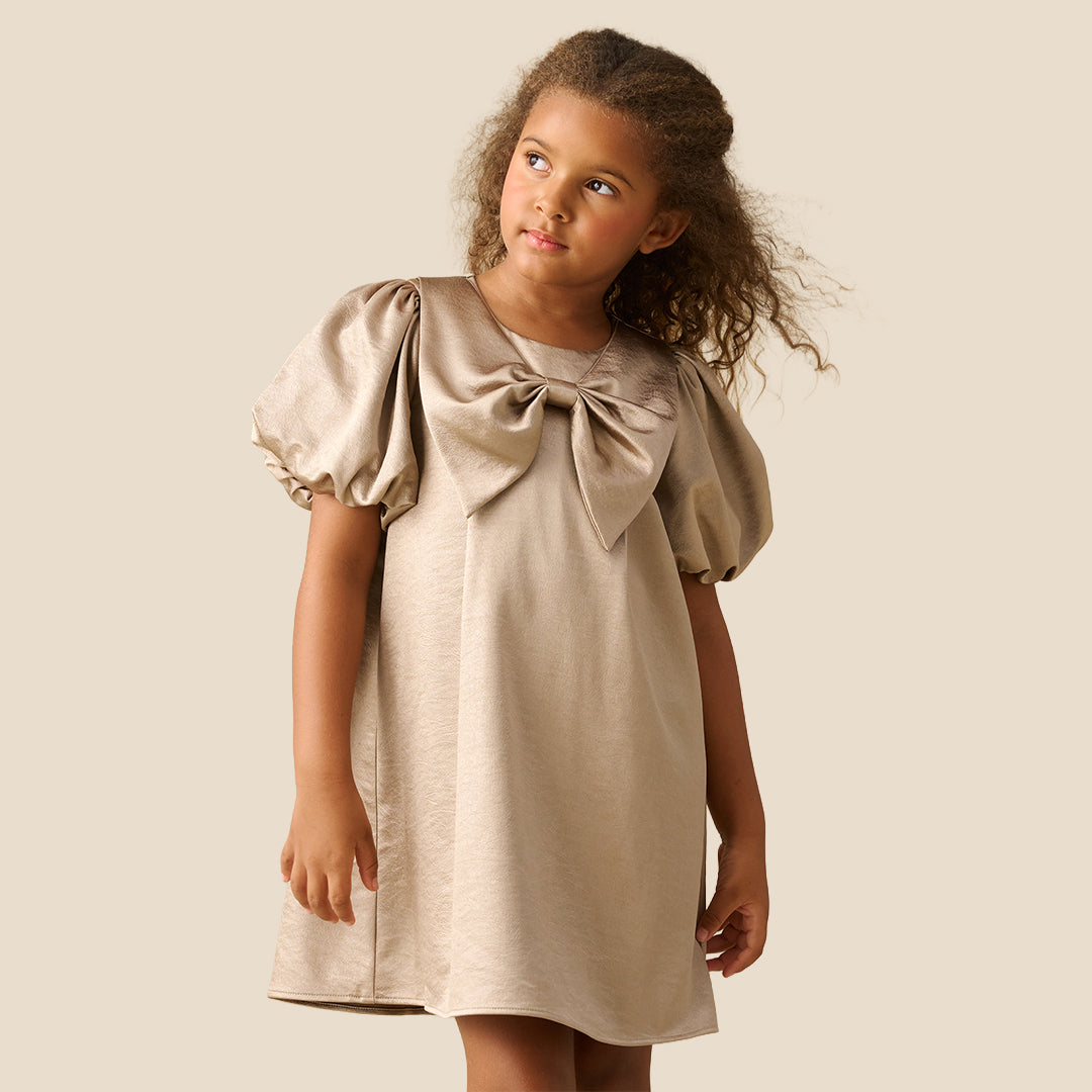 A young girl with curly brown hair wearing a champagne beige dress with puffed short sleeves and a large bow at the neckline, standing against a solid light beige background.