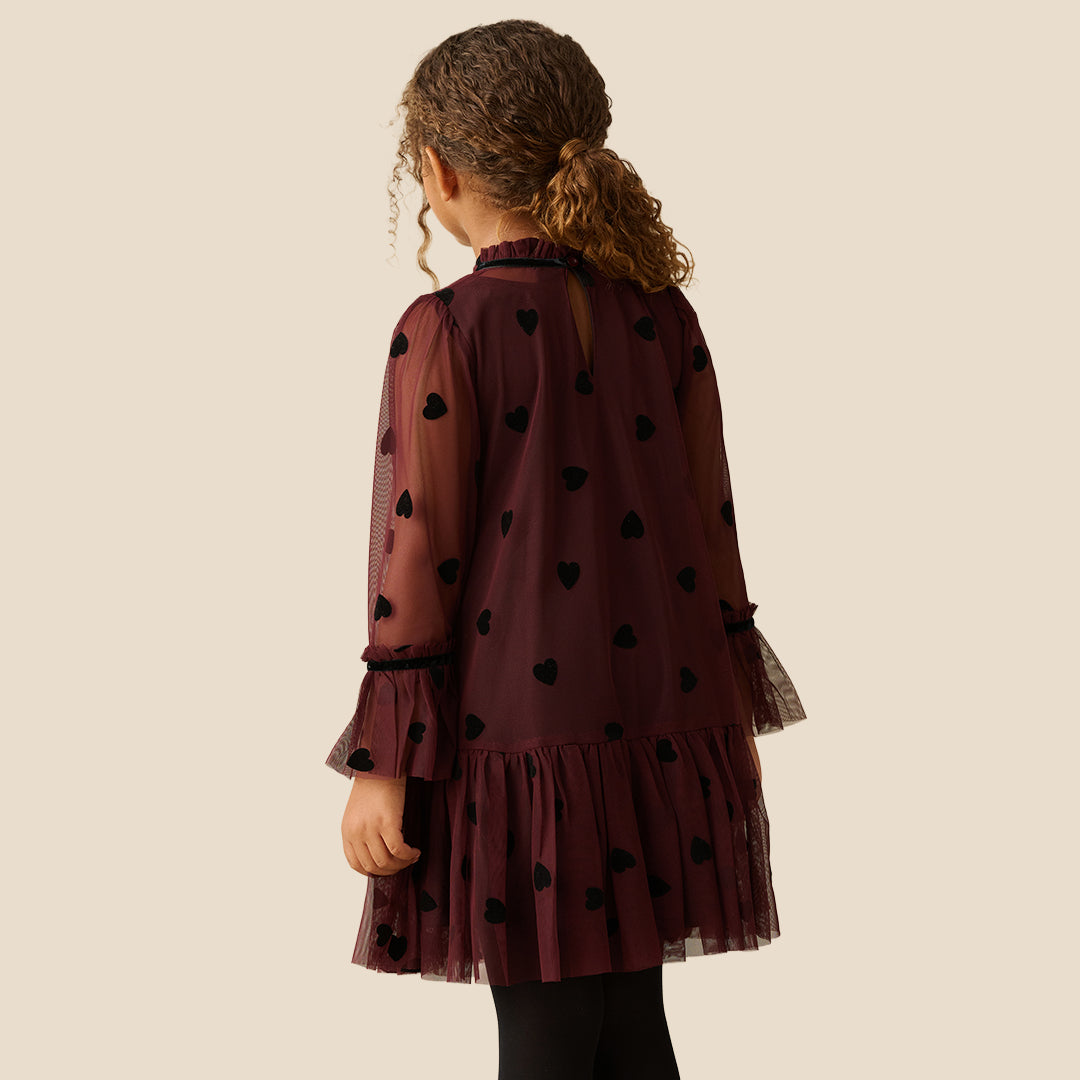 A young girl with curly brown hair wearing a burgundy sheer mesh dress with black heart patterns and a ruffled tulle hem, standing against a solid beige background.