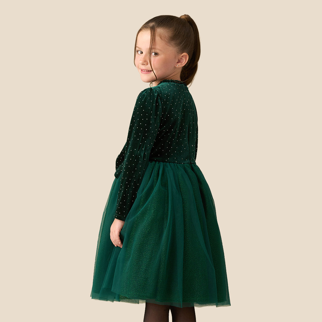 A young girl wearing a dark green velvet long-sleeve dress with a polka-dot pattern, a matching tulle skirt, and a large bow at the waist, standing against a solid beige background.