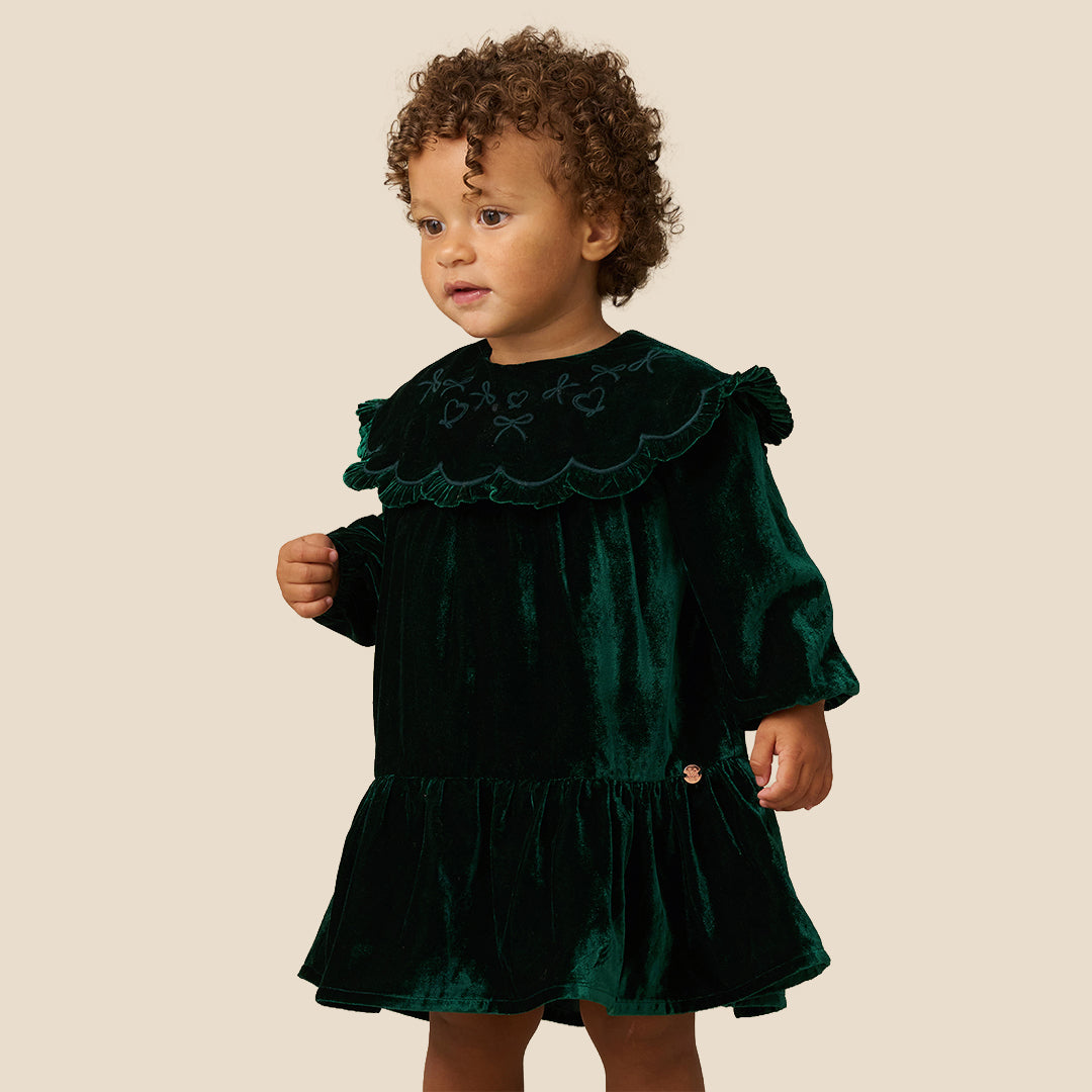 Two young children standing barefoot against a plain beige studio background, wearing matching dark green velvet dresses with wide ruffled shoulders and long sleeves.
