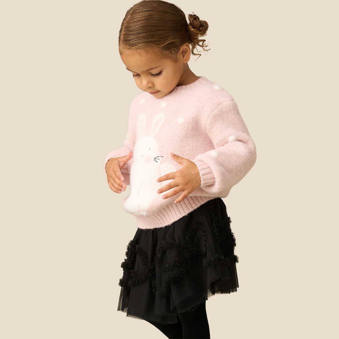 A young girl smiling while wearing a light pink knit sweater with a white bunny applique and white pom-pom details, paired with a black ruffled tutu skirt, against a solid beige background.